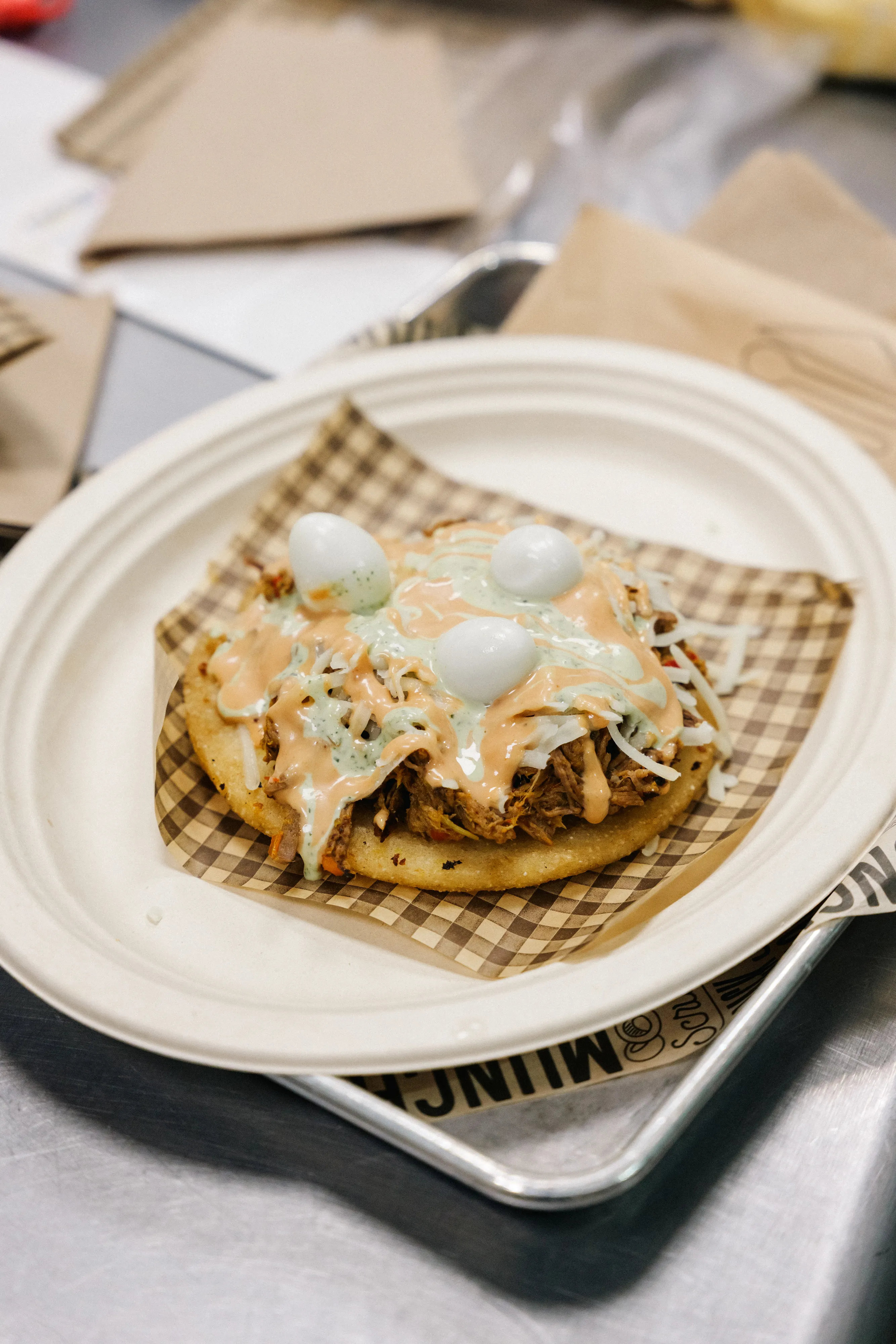 An arepa topped with shredded chicken, melted cheese and quail eggs, served on a paper plate